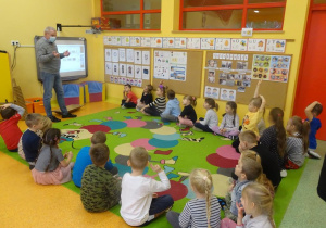 Pan Maciej Grzeszczak przedstawia dzieciom siedzącym przed tablicą interaktywną prezentację dotyczącą dawnych zamierających zawodów.